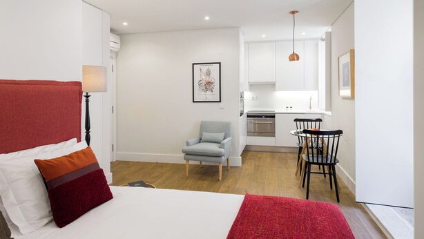 A bedroom with a bed, a chair, and a kitchenette in the background. - LSA Bairro Alto by Numa (Lisbon)