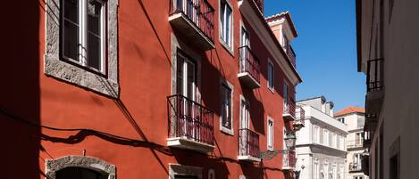 Une rue étroite bordée d’immeubles aux façades rouges et blanches, de balcons et surplombée d’un ciel d’un bleu limpide.