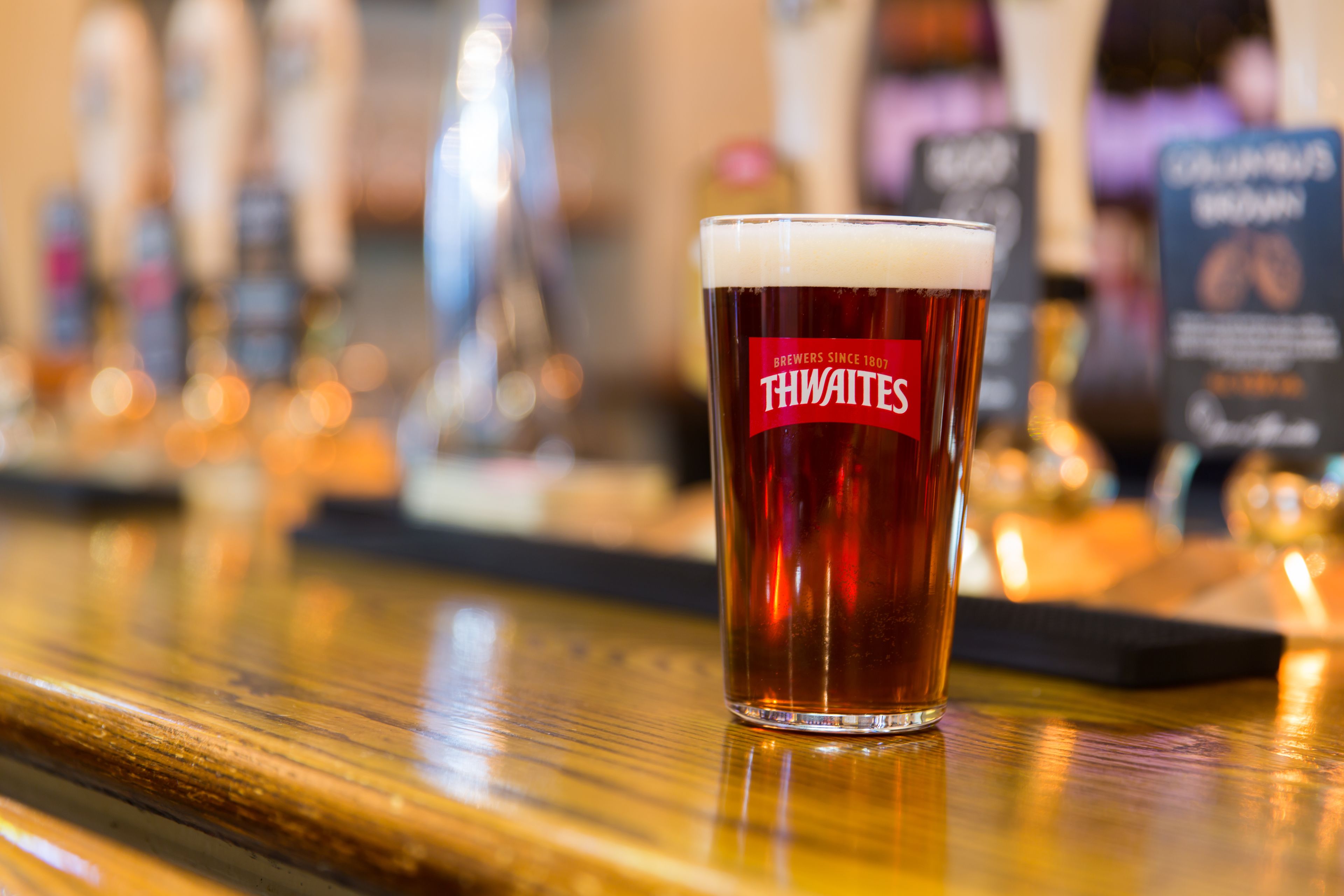 Un verre de bière avec une étiquette portant l’inscription « THWAITES » sur le comptoir d’un bar.
