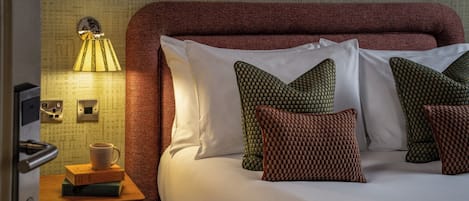 A hotel room with a large bed, multiple pillows, a bedside lamp, a wooden nightstand, and a wall with a patterned design.