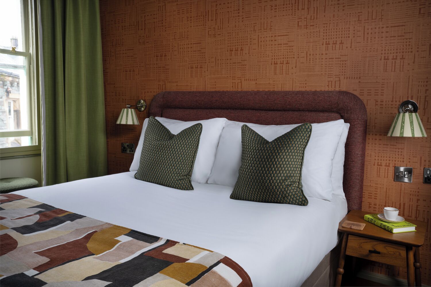 A hotel room with a large bed, two pillows, a patterned throw, a nightstand with a lamp, and a window with curtains.