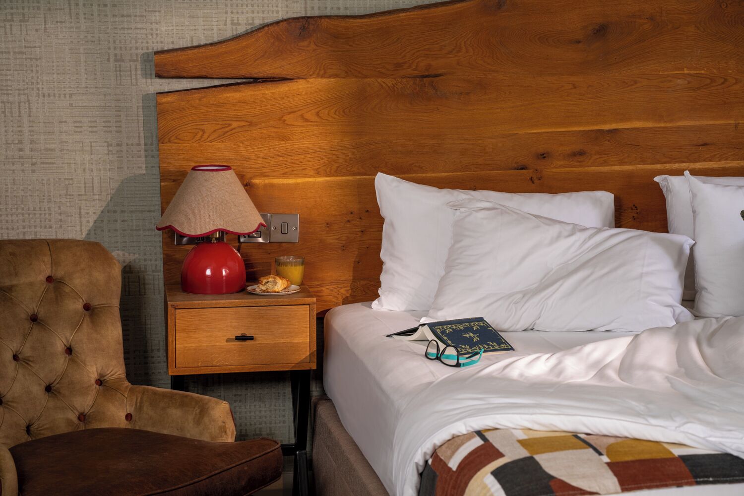 A hotel room with a wooden headboard, a bedside table with a lamp, a chair, and a bed with pillows and a book.