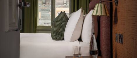 A hotel room with a bed, pillows, a bedside table with a glass, a bottle, and a lamp.