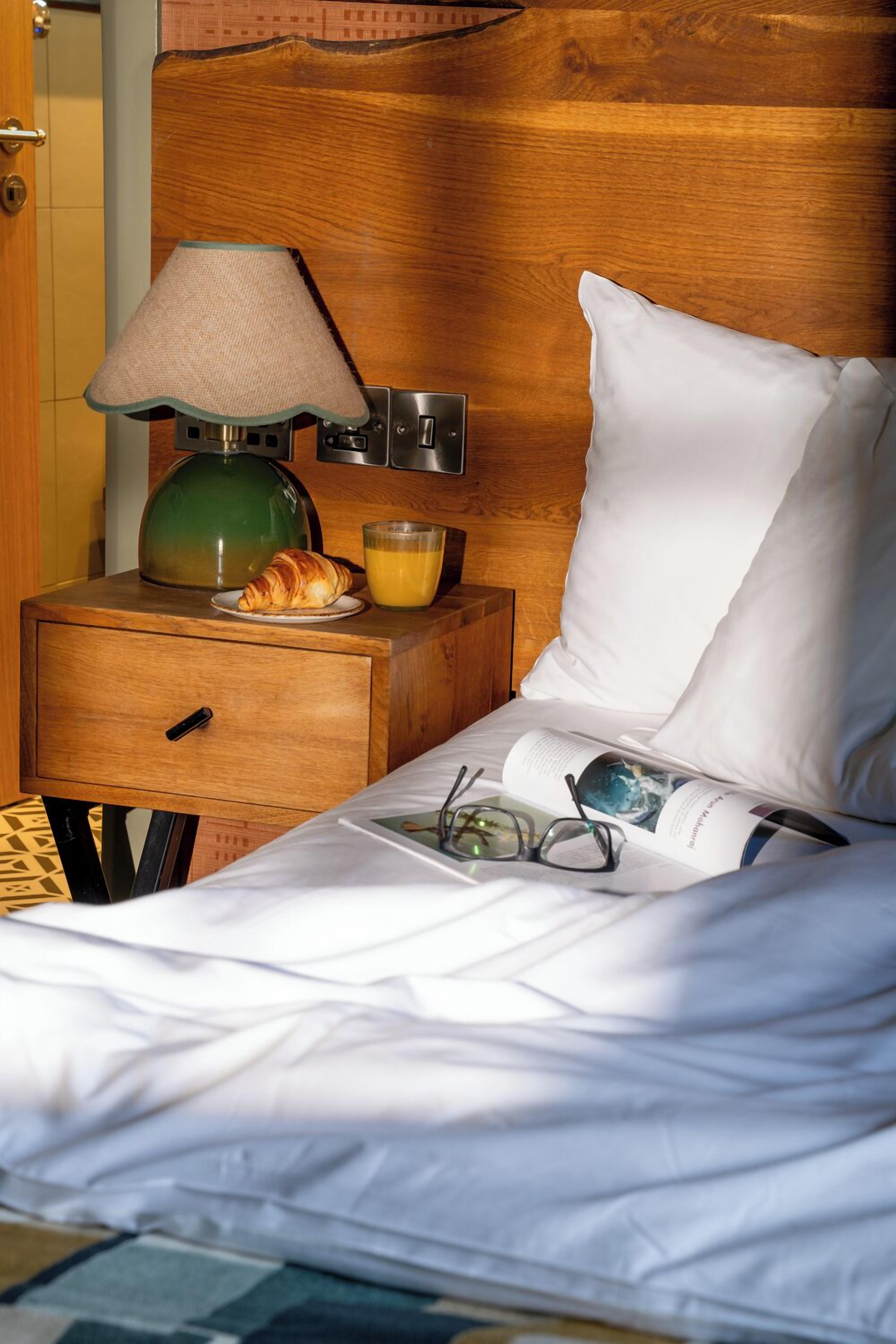 Un lit bien fait, avec du linge de lit blanc, une lampe verte, une assiette contenant un croissant et du jus d’orange, et un magazine sur la table de chevet.