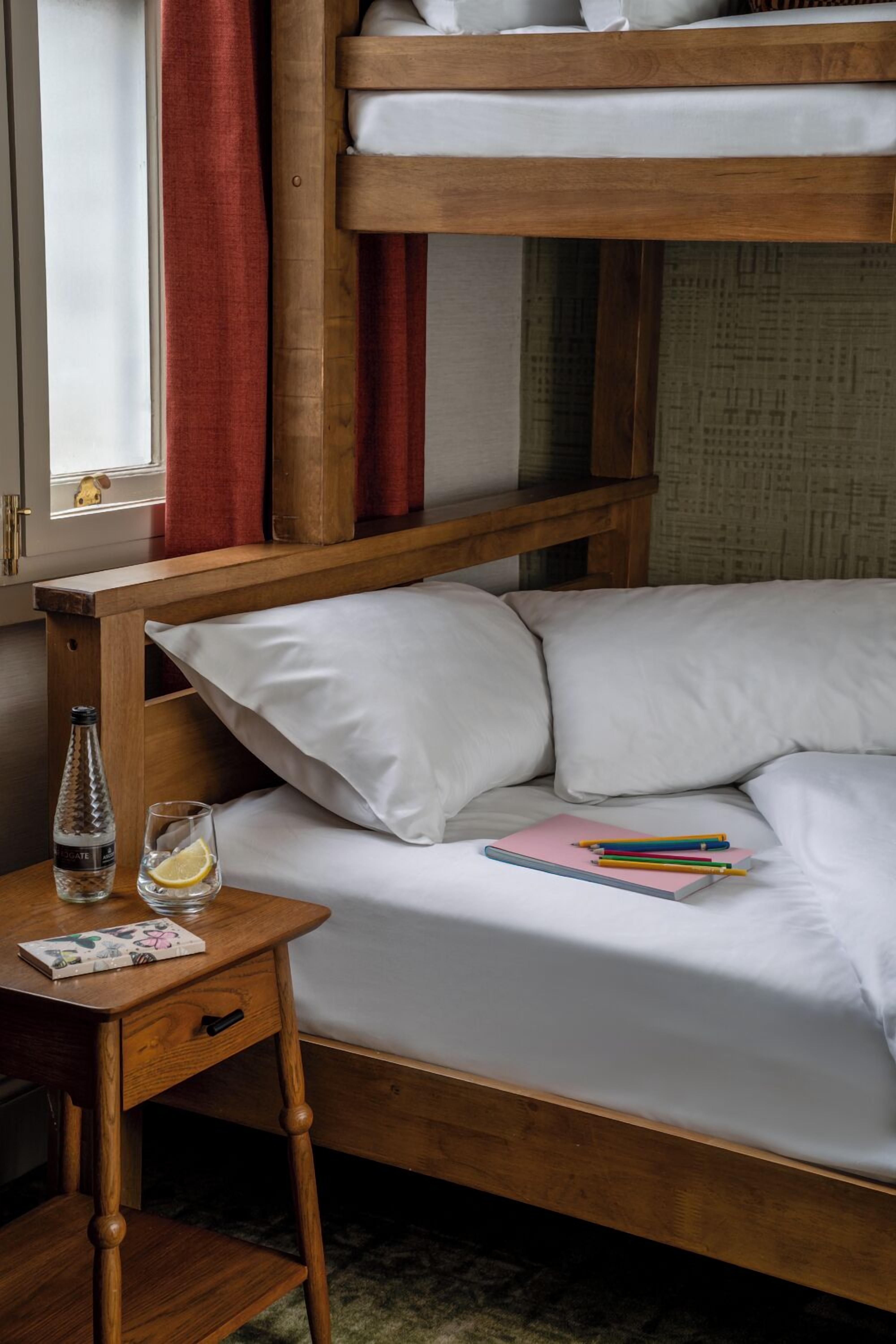 A bunk bed with white bedding, a wooden nightstand with a bottle and glasses, and a pink notebook.