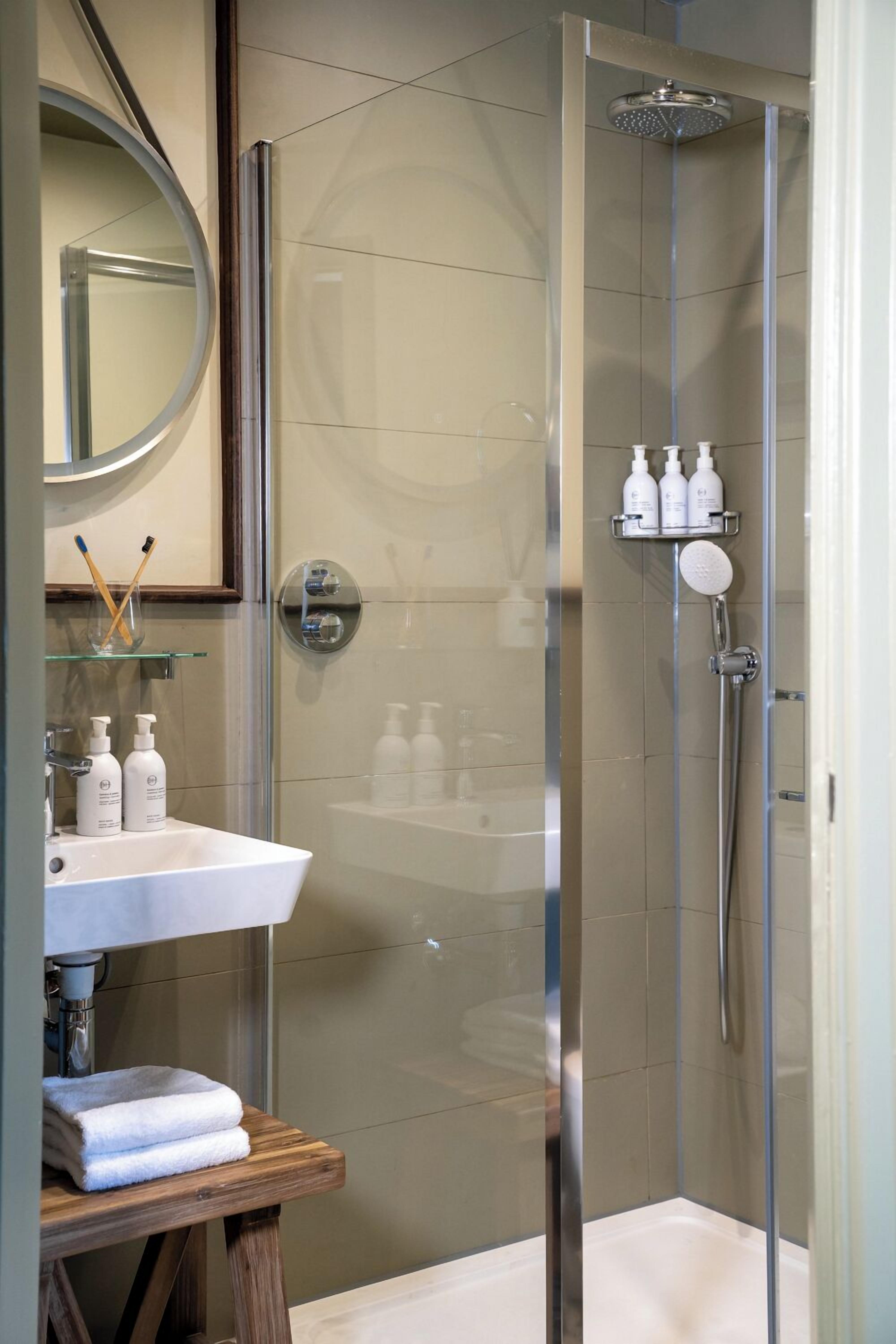 A modern bathroom with a glass-enclosed shower, a round mirror, and a wooden stool with towels.