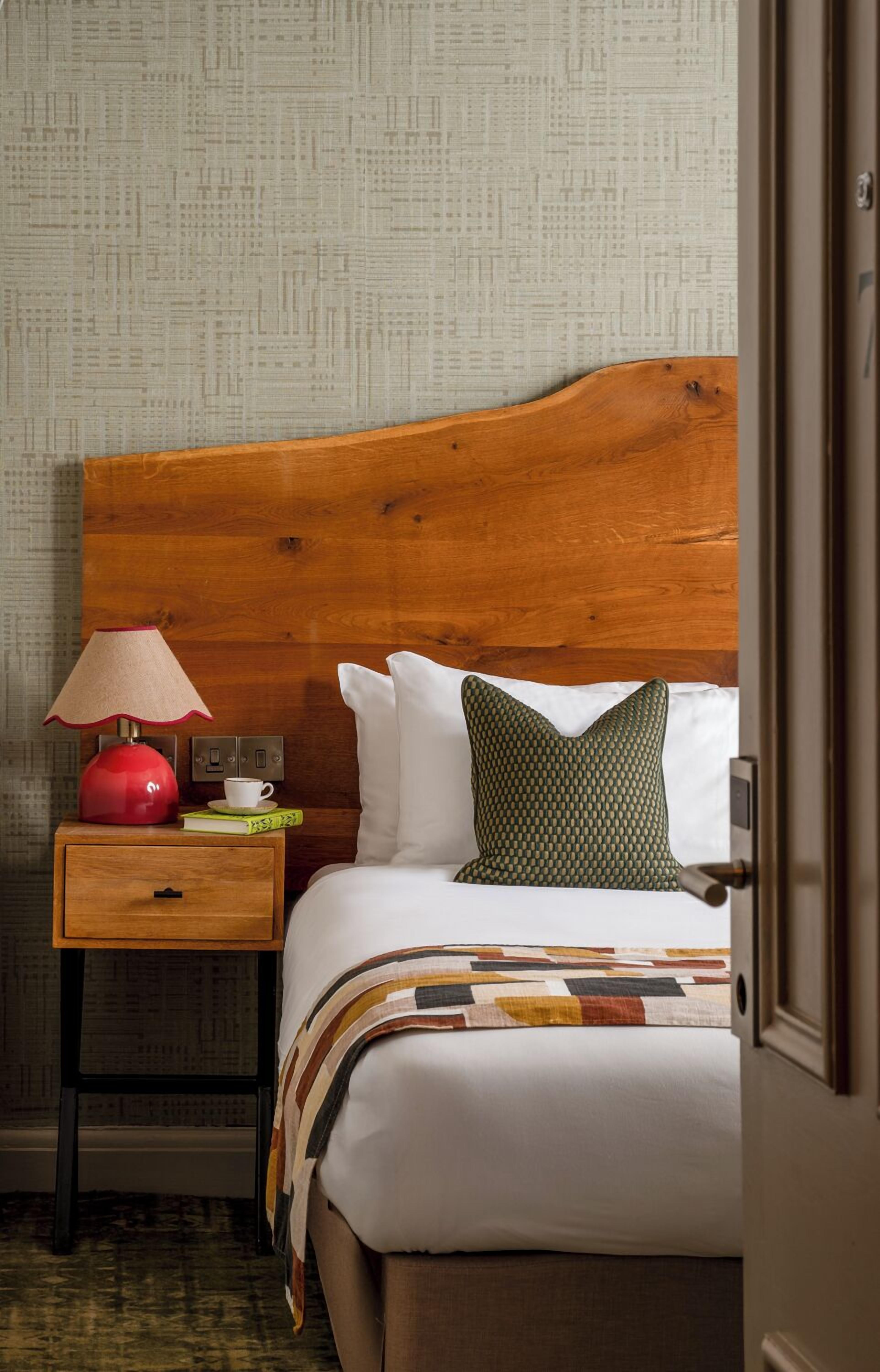 A hotel room with a wooden headboard, a bedside table with a lamp, a quilted pillow, and a book.