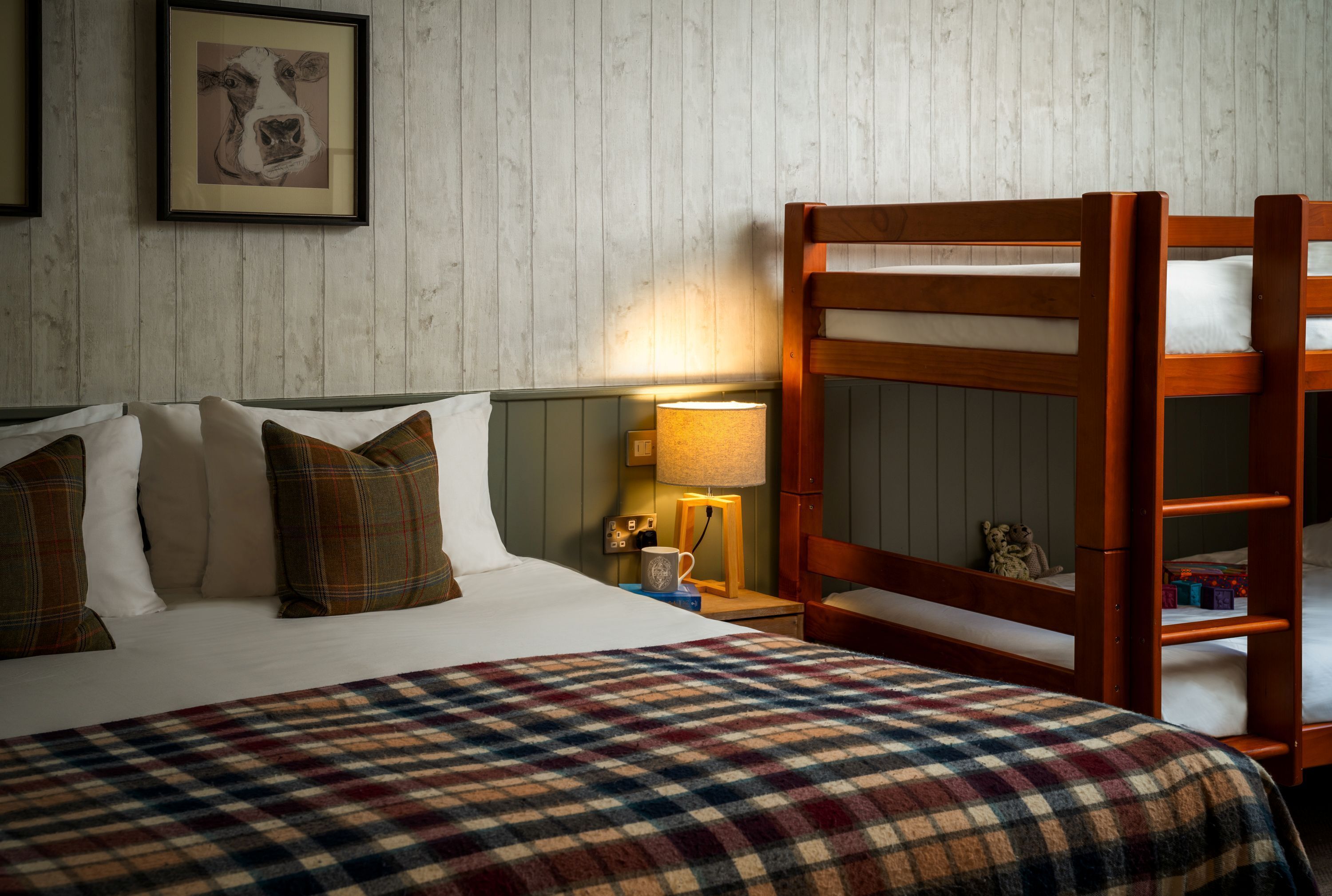 A bedroom with a bed, pillows, a plaid blanket, a wooden bunk bed, a nightstand with a lamp, and a framed picture on the wall.