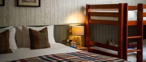 A bedroom with a bed, pillows, a plaid blanket, a wooden bunk bed, a nightstand with a lamp, and a framed picture on the wall.