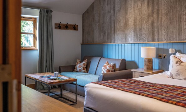 Una habitación de hotel con cama, sofá, mesa con lámpara y ventana con cortinas.