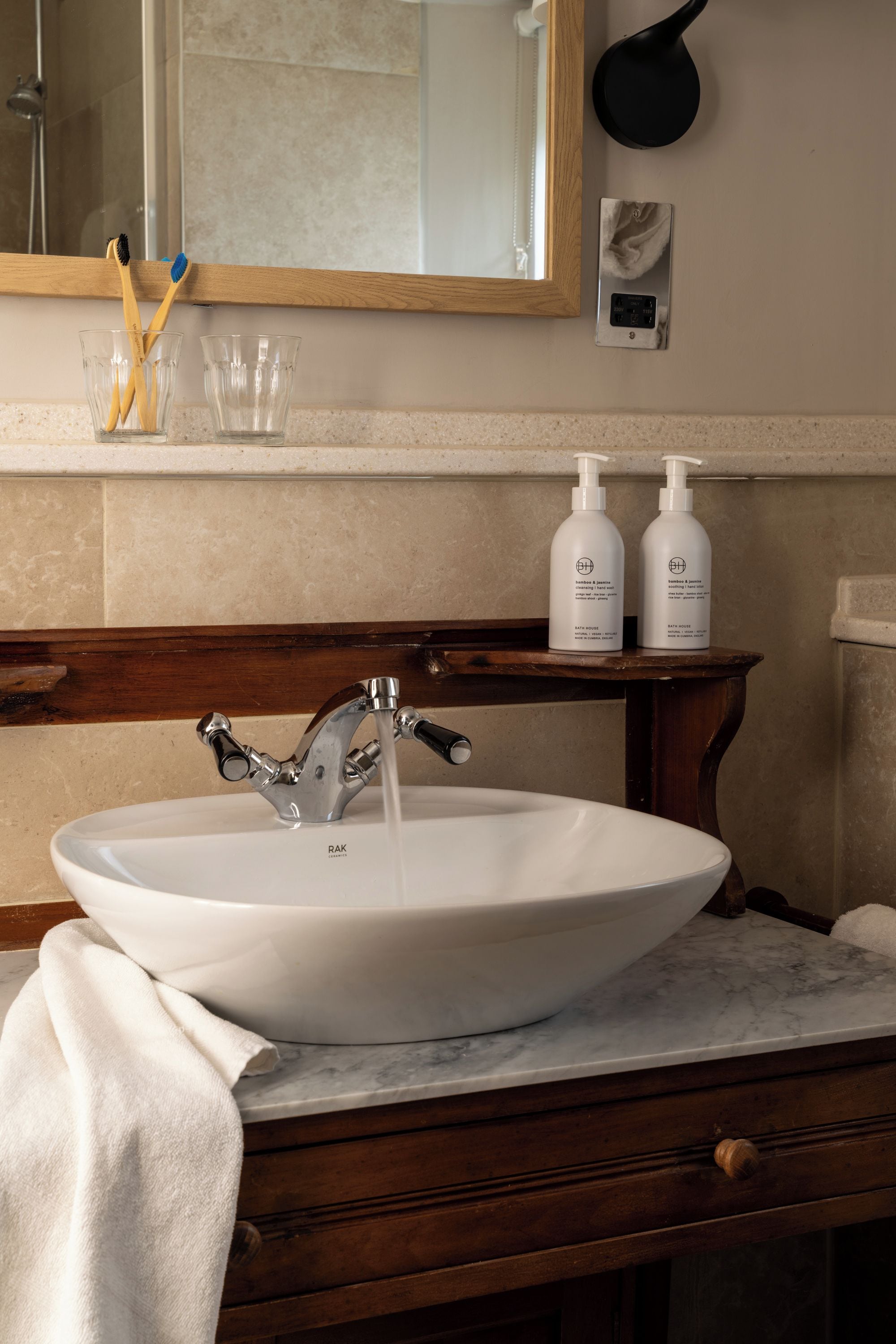 Un baño con un lavabo de cerámica blanca, un mueble de tocador de madera, un espejo y dos botellas de champú natural de casa de baños.