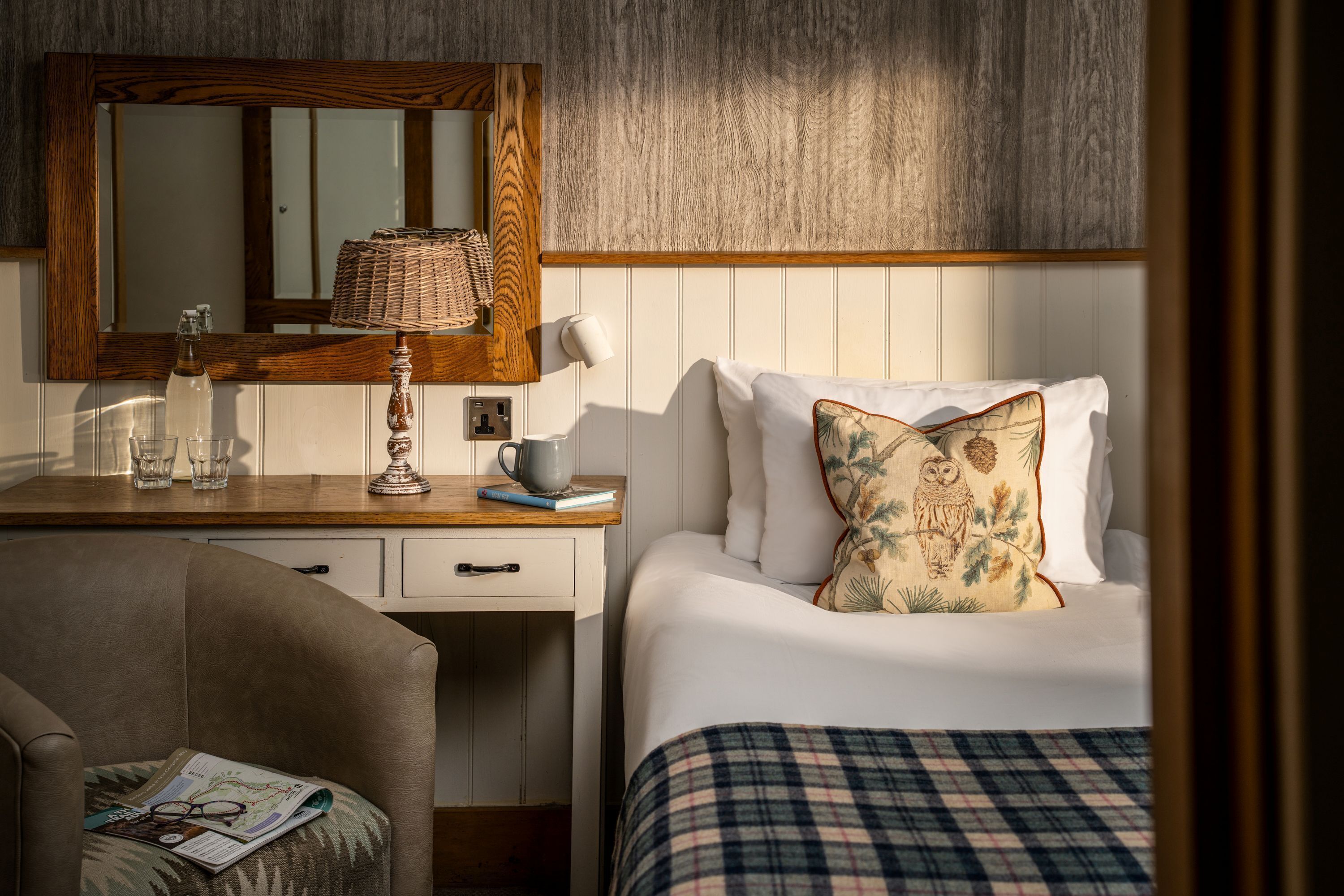 Un dormitorio ordenado con cabecera de madera, mesita de noche con lámpara, espejo y cama con manta escocesa y cojín decorativo.
