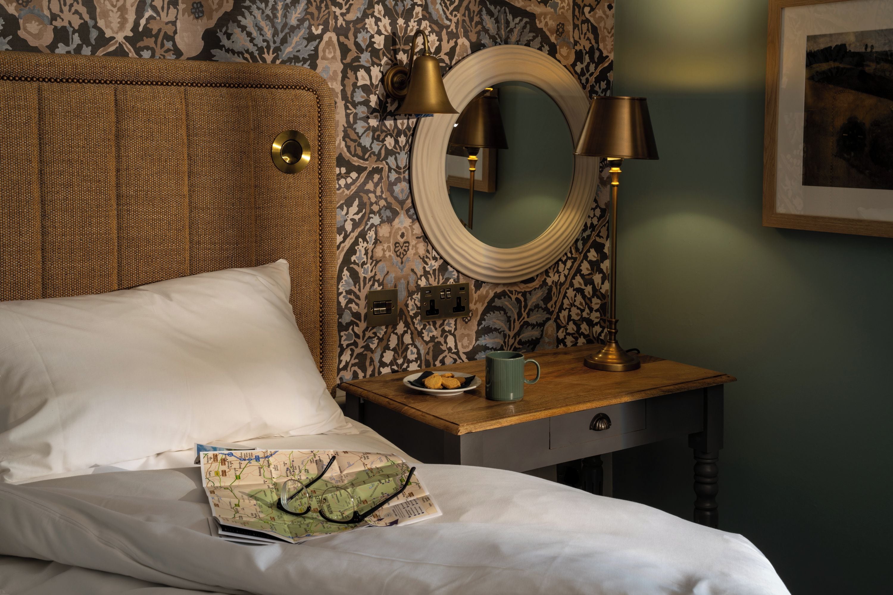 A neatly made bed with white linens, a wooden bedside table with a plate of cookies, a green mug, and a map.