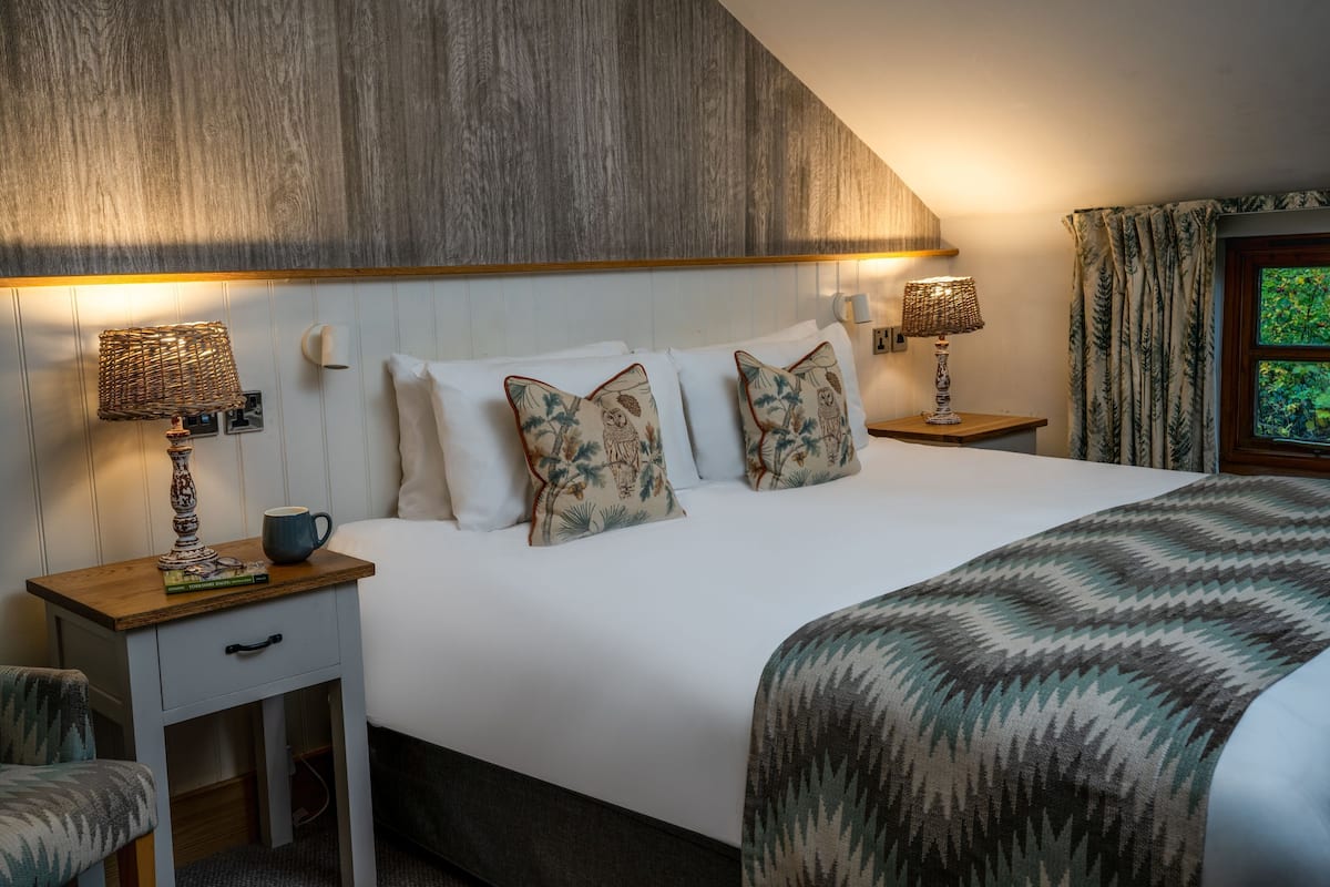 A bedroom with a wooden headboard, two bedside tables with lamps, a bed with patterned pillows, a bedside drawer, and a window with curtains.