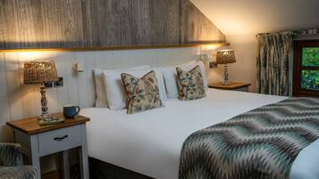 Quarto com cabeceira de madeira, duas mesas de cabeceira com abajures, cama com almofadas estampadas, gaveta lateral e janela com cortinas.
