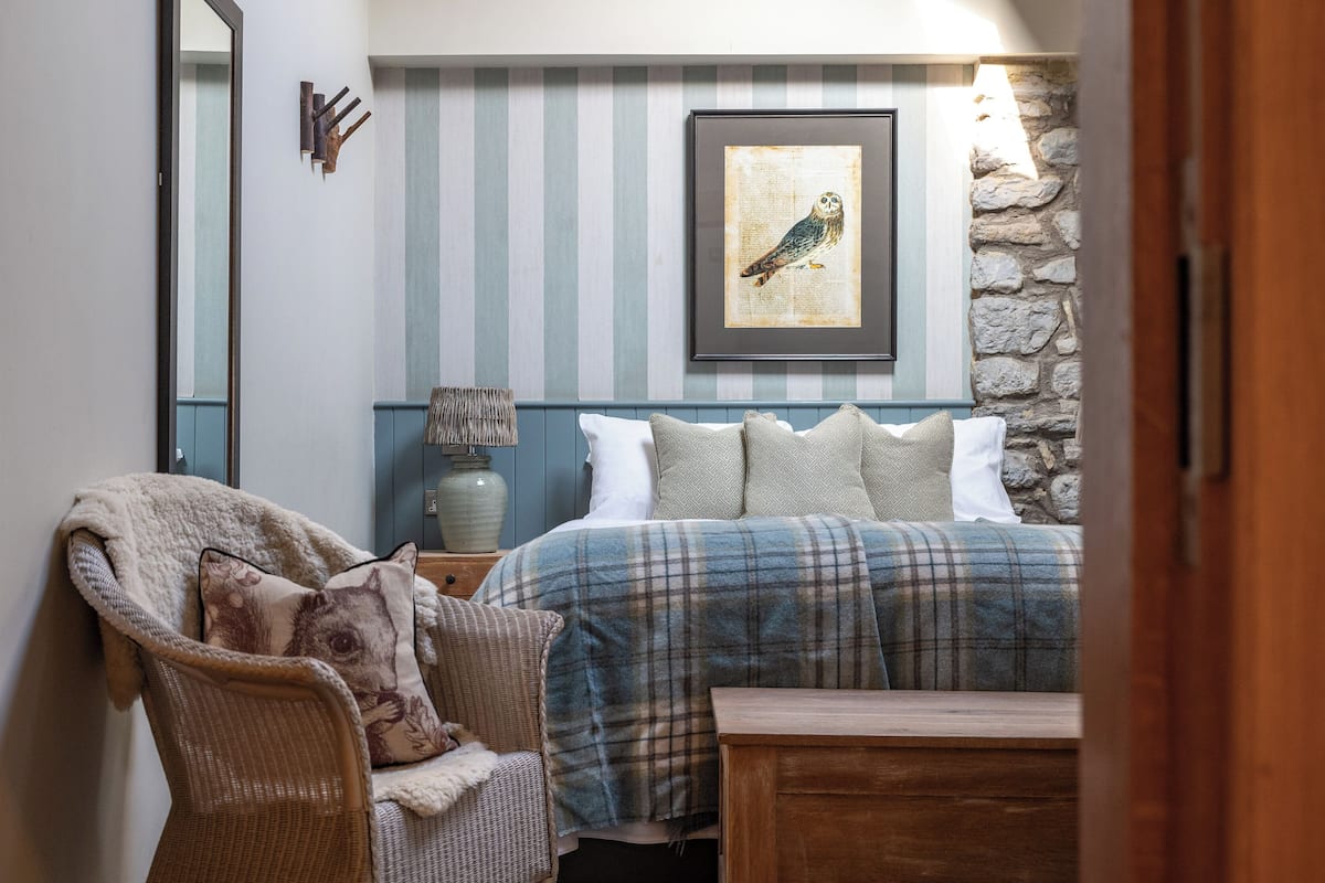 A cozy bedroom with a plaid bedspread, a wicker chair with cushions, a bedside table with a lamp, and a framed owl artwork on the wall.
