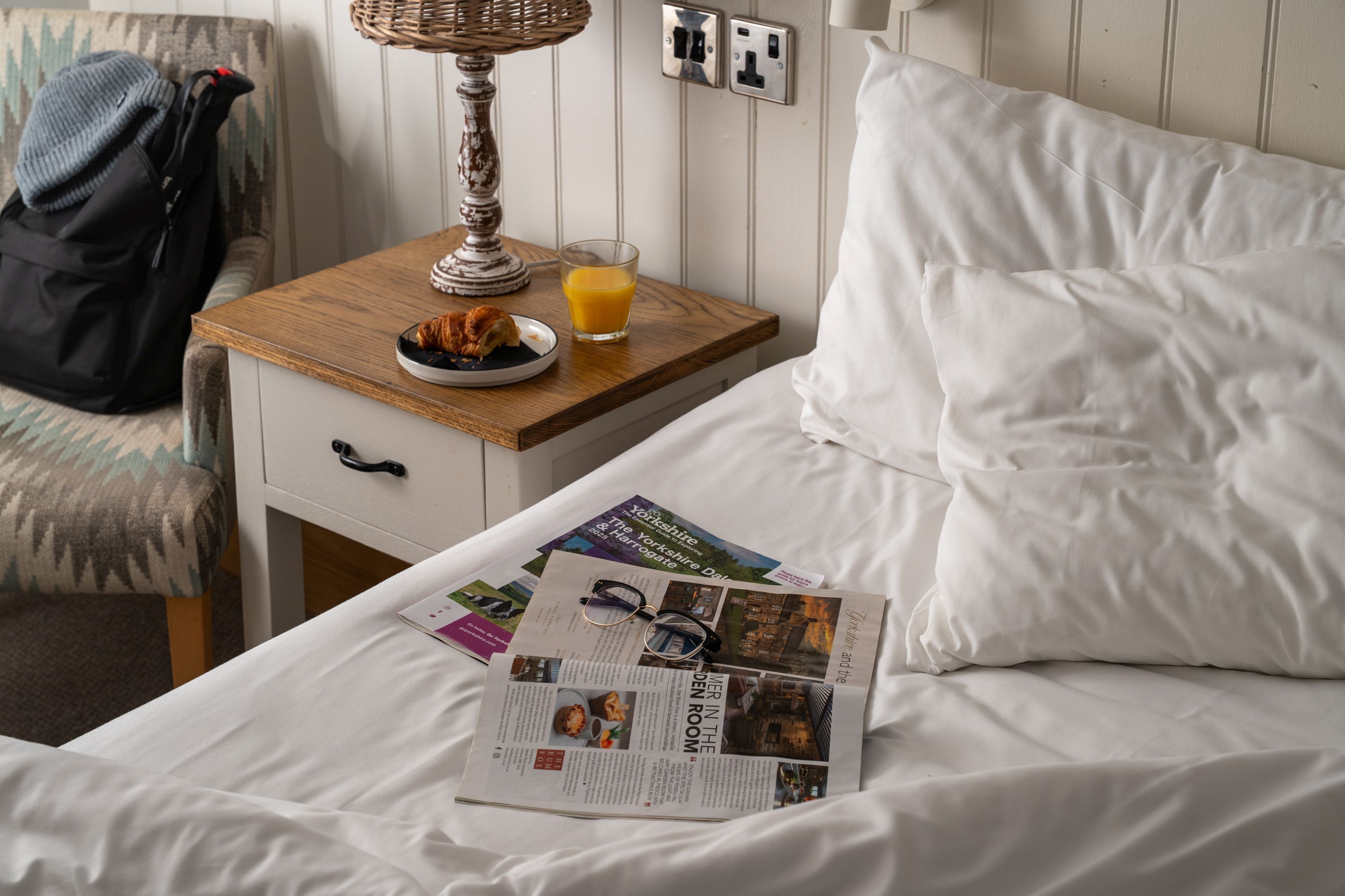 A neatly made bed with white linens, a bedside table with a lamp, a glass of orange juice, and a magazine on the bed.