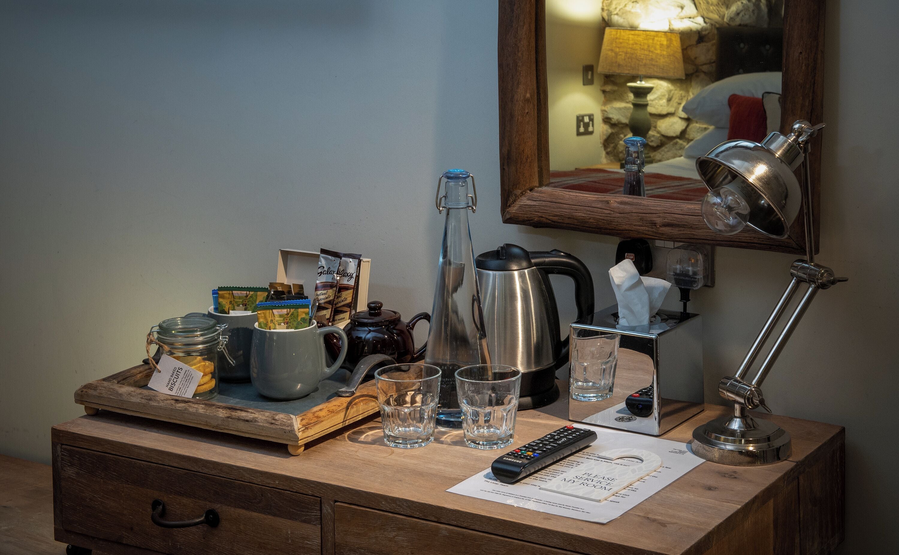 Una mesa de madera con una cafetera, un hervidor, un control remoto, un vaso de agua y una bandeja con frascos y paquetes.