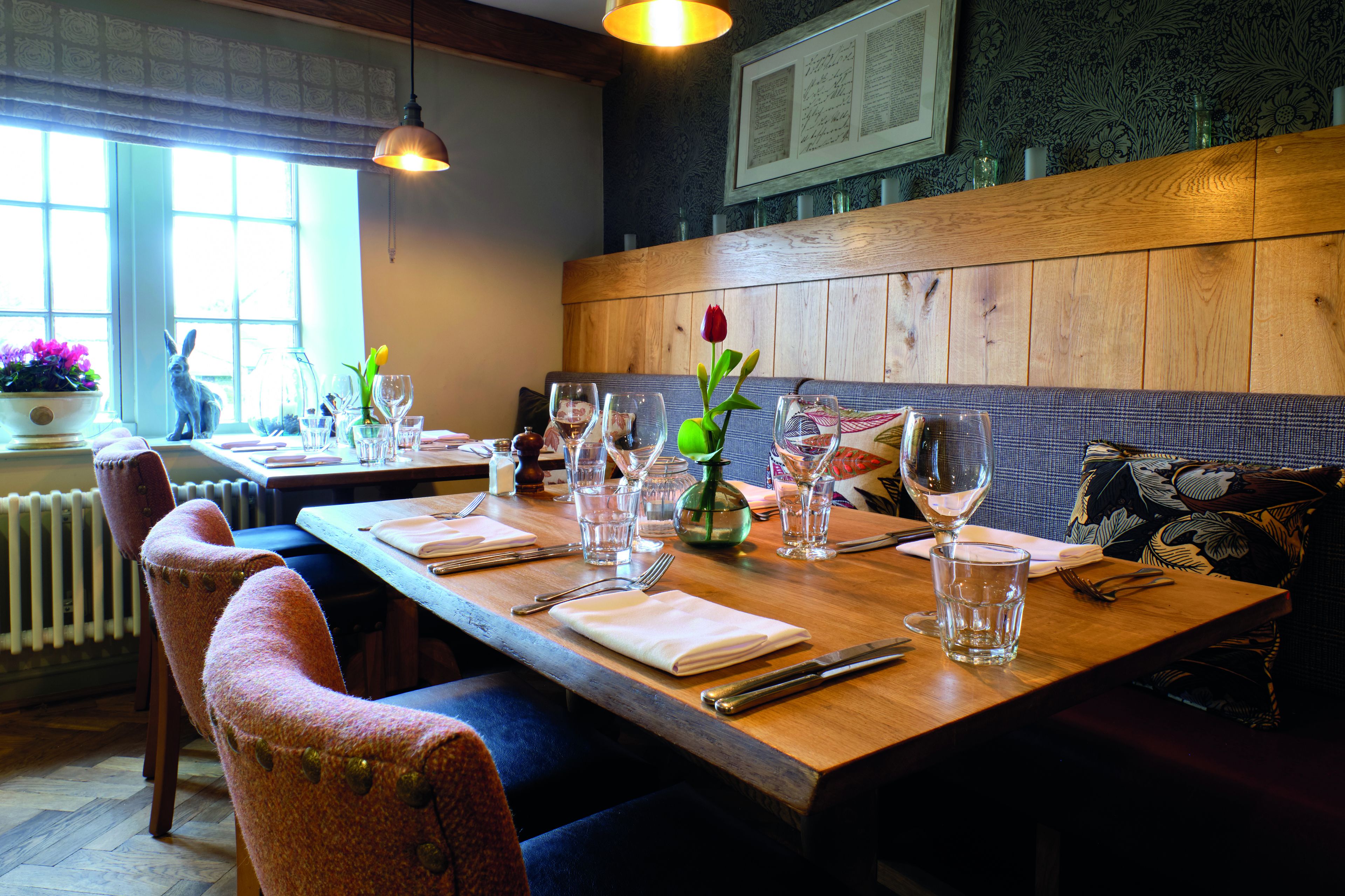 Un restaurante con una mesa de madera lista para comer, sillas y un jarrón con flores.