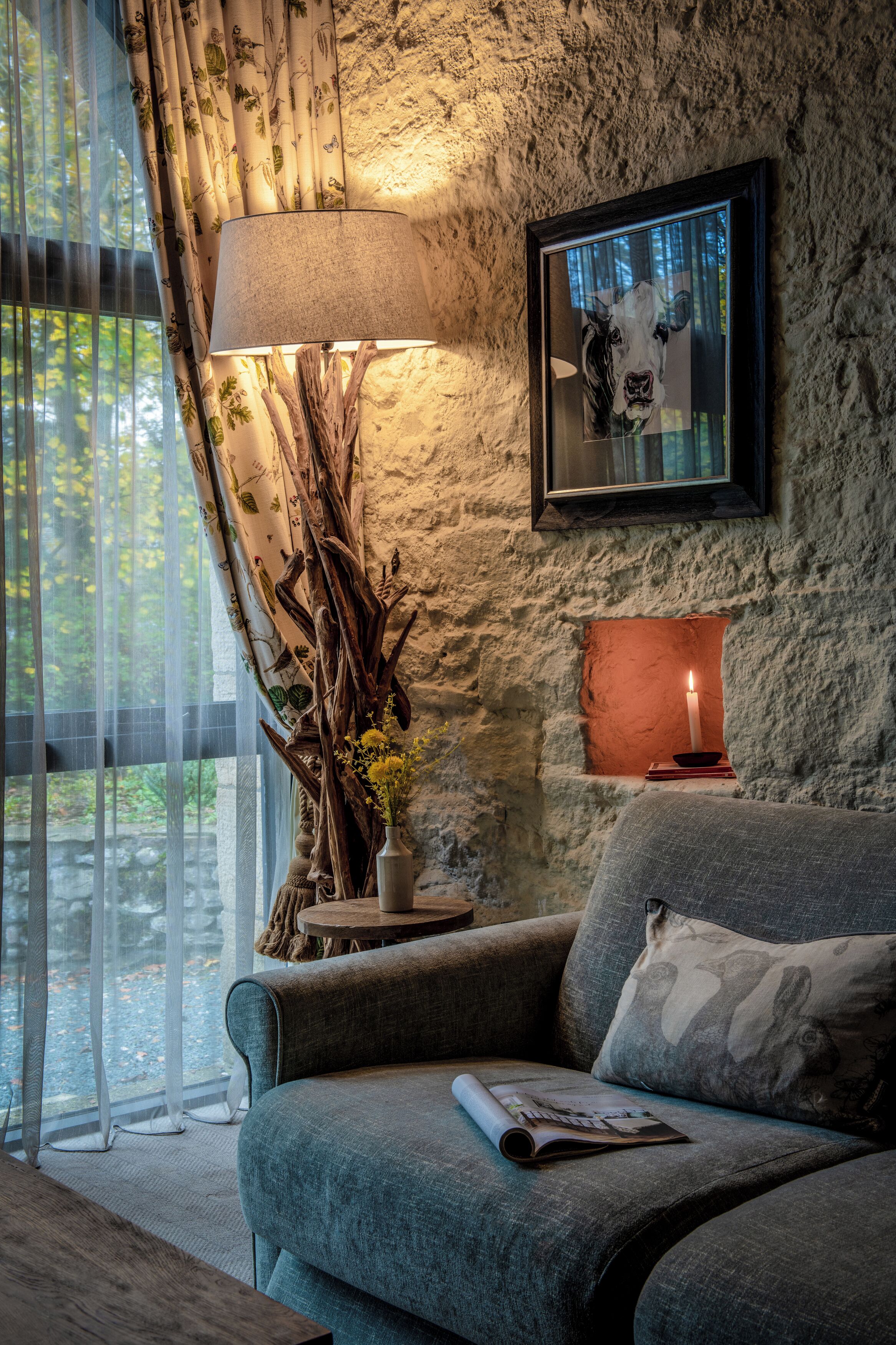 Ein gemütliches Wohnzimmer mit Steinwand, einem gerahmten Kunstwerk, einer Lampe und einem Fenster mit leichtem Vorhang.