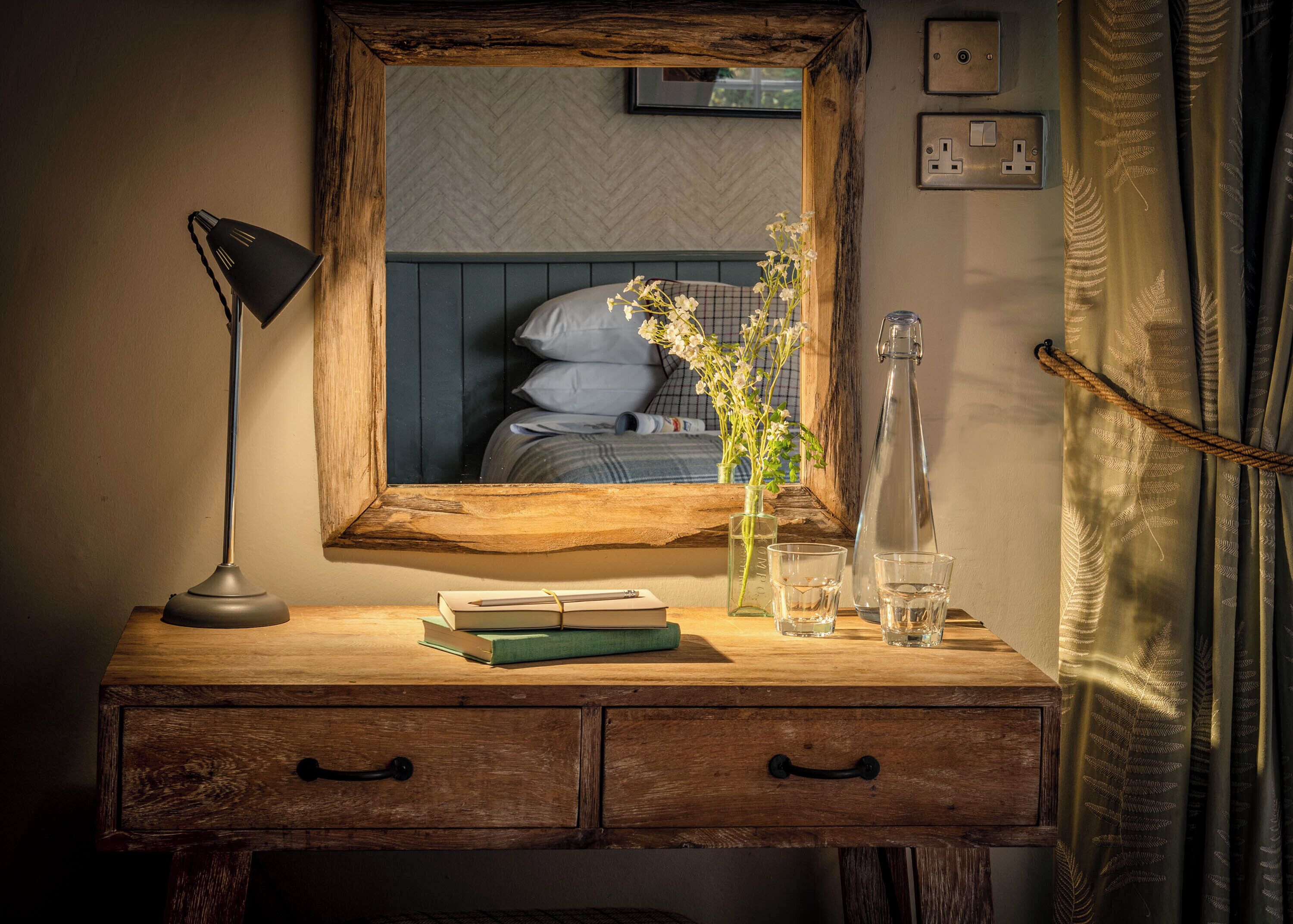 Un escritorio de madera con un espejo, libros, una lámpara y un jarrón con flores.