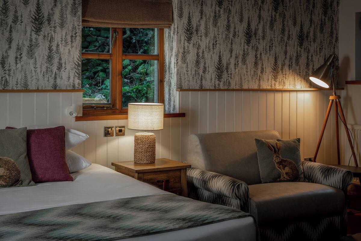 A bedroom with a bed, two chairs, a nightstand with a lamp, and a window with a view of greenery.