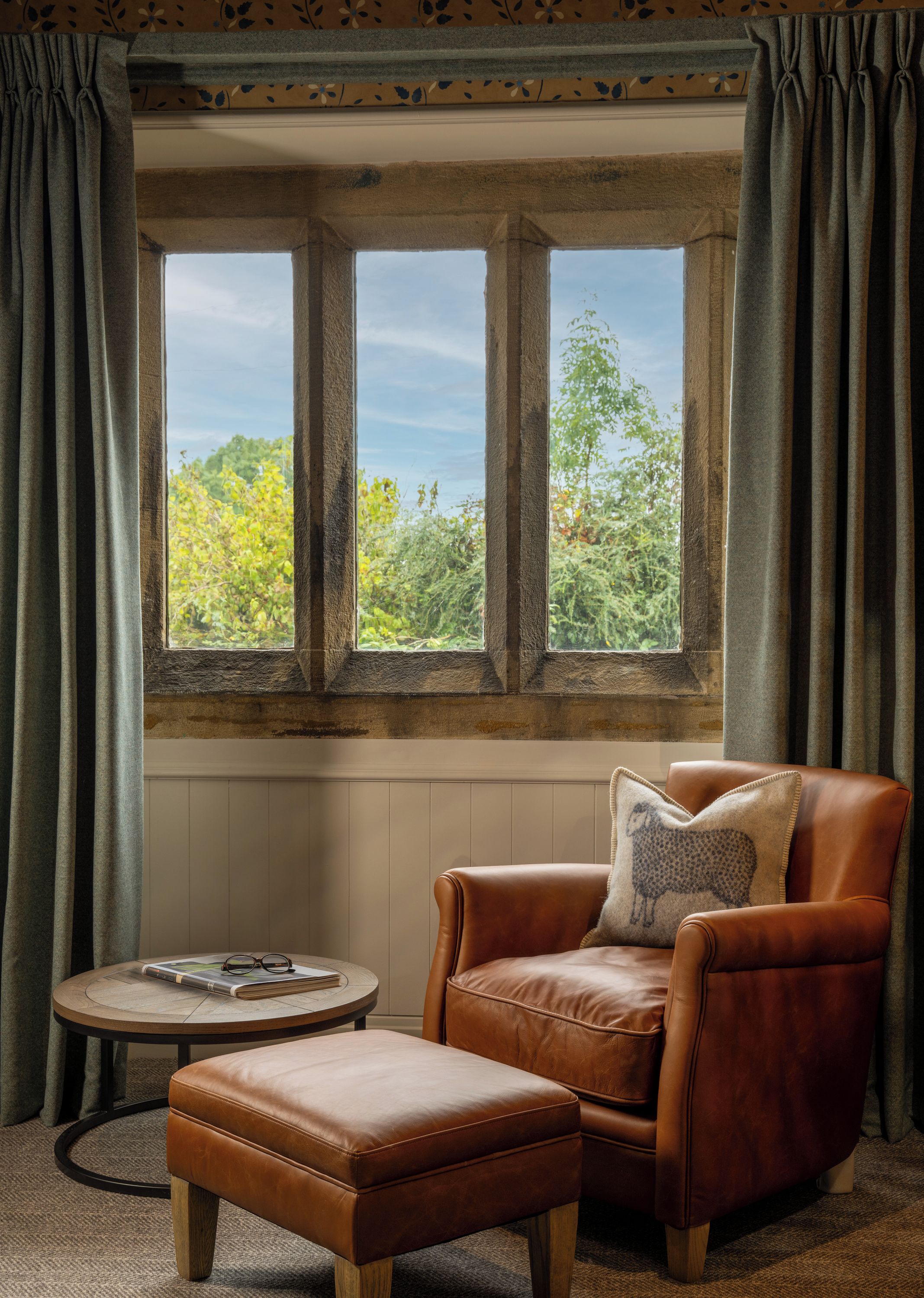 A room with a large window, a leather armchair, a small round table, and a matching ottoman.