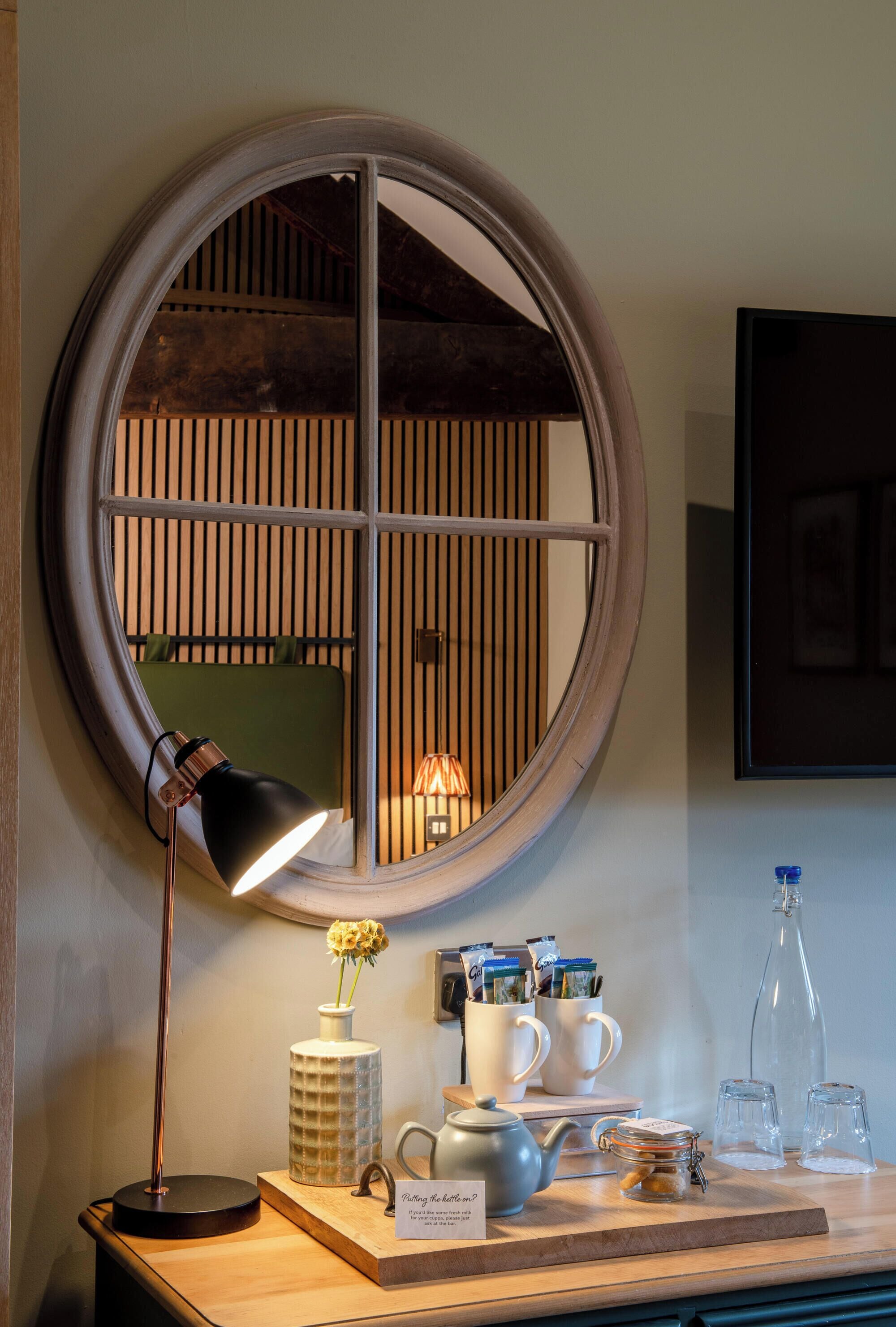 A round mirror with a wooden frame, a desk with a lamp, a vase with flowers, a teapot, and a mug set.