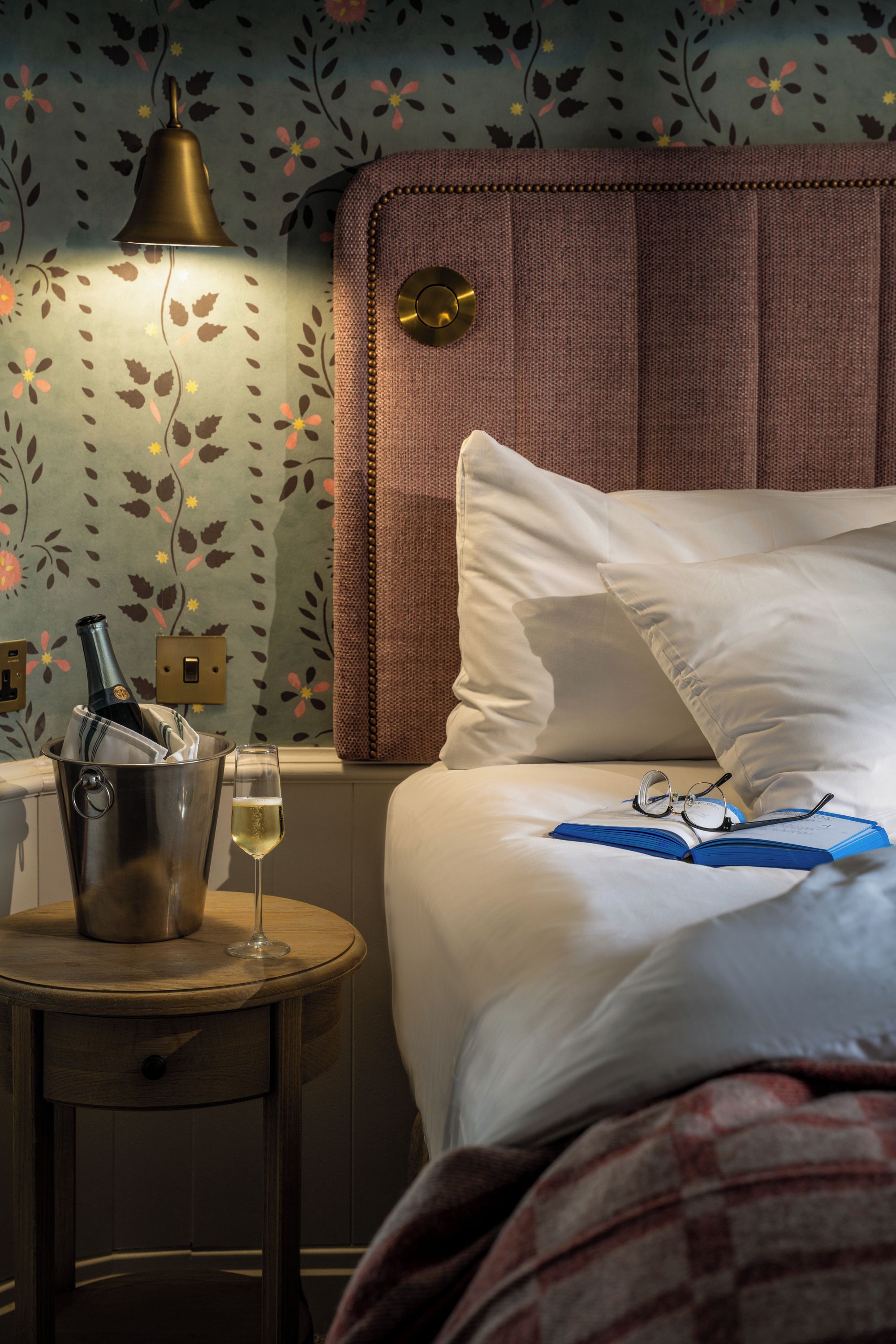 Una habitación de hotel con cama, mesita de noche, lámpara, una botella de champán y copas.