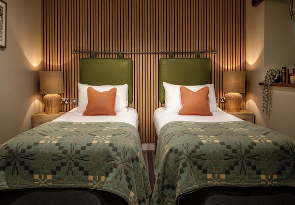 A double room with two beds, each with a green headboard, white bedding, and patterned green blankets. There are two bedside tables with lamps and a small plant on the right side. - Lister Arms (Skipton)