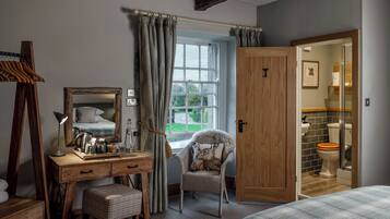 A bedroom with a wooden desk, a chair with a cushion, a bed, a window with curtains, and a bathroom visible through an open door.