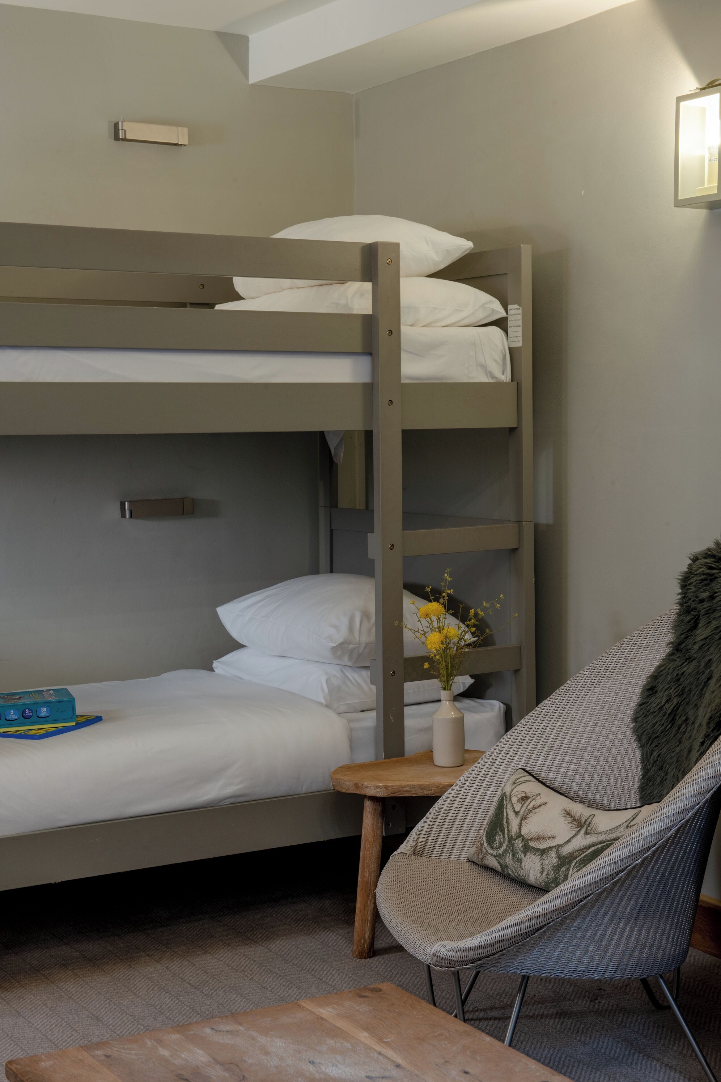 Een stapelbed met witte lakens, een kleine tafel met een bloemenvaas en een fauteuil met een vacht.'