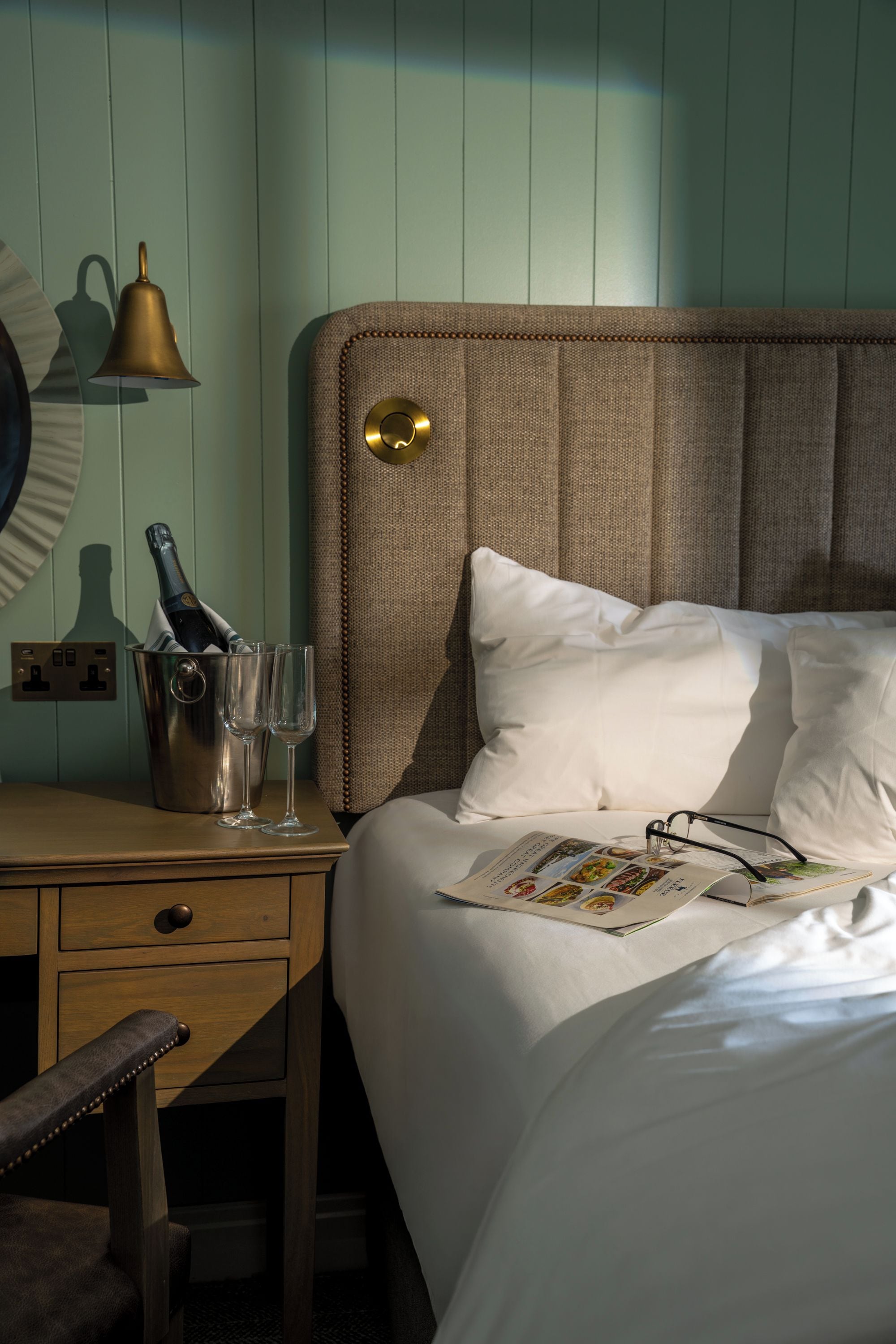 A neatly made bed with pillows, a bedside table with a bottle and glasses, a lamp, and a wall-mounted clock.