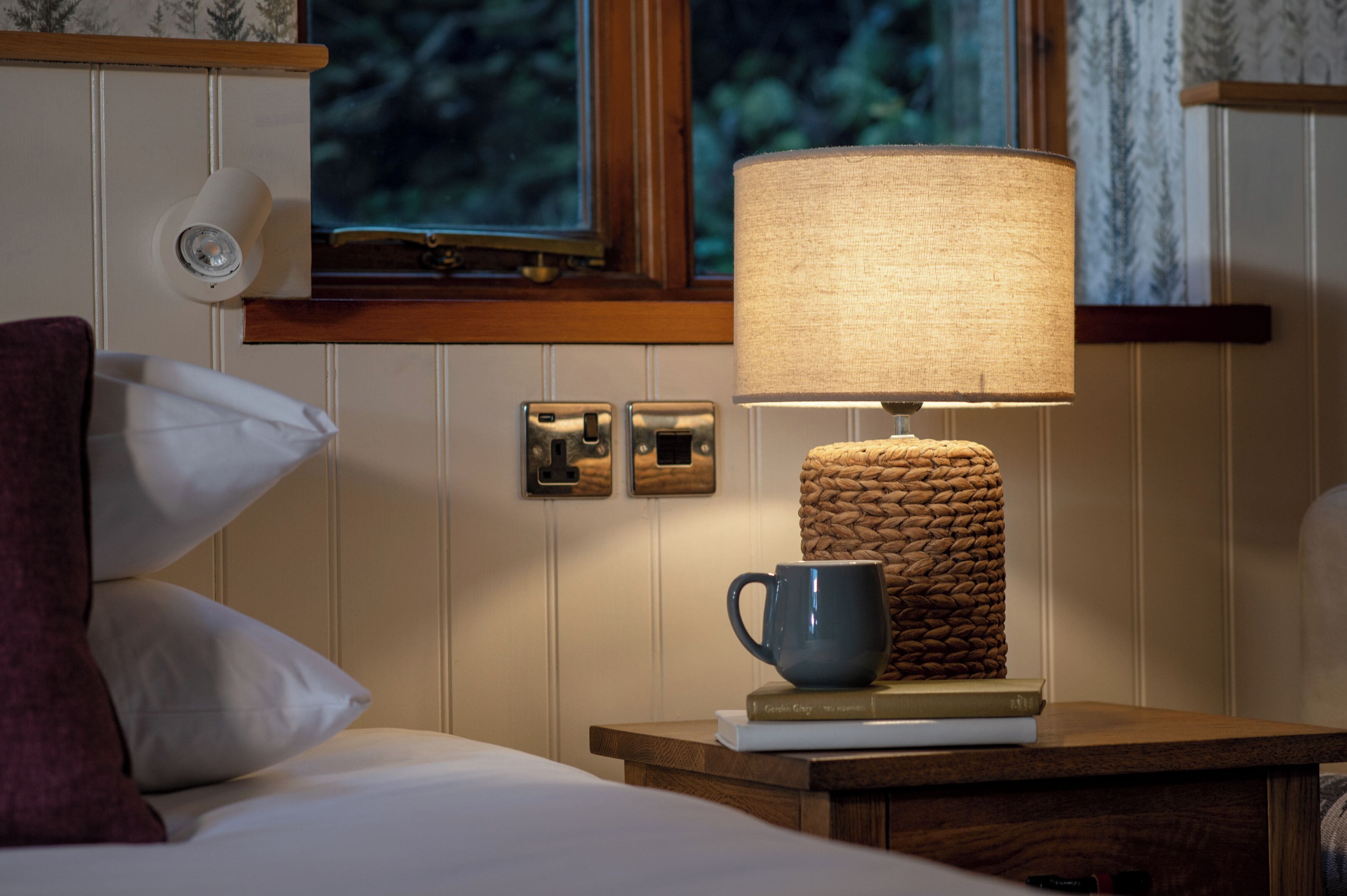 Una cama bien hecha con almohadas blancas y púrpuras, una mesita de noche con una lámpara, una taza y libros.