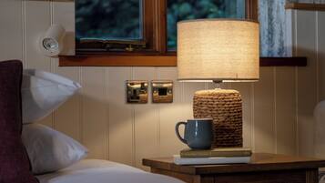 Una cama bien hecha con almohadas blancas y púrpuras, una mesita de noche con una lámpara, una taza y libros.