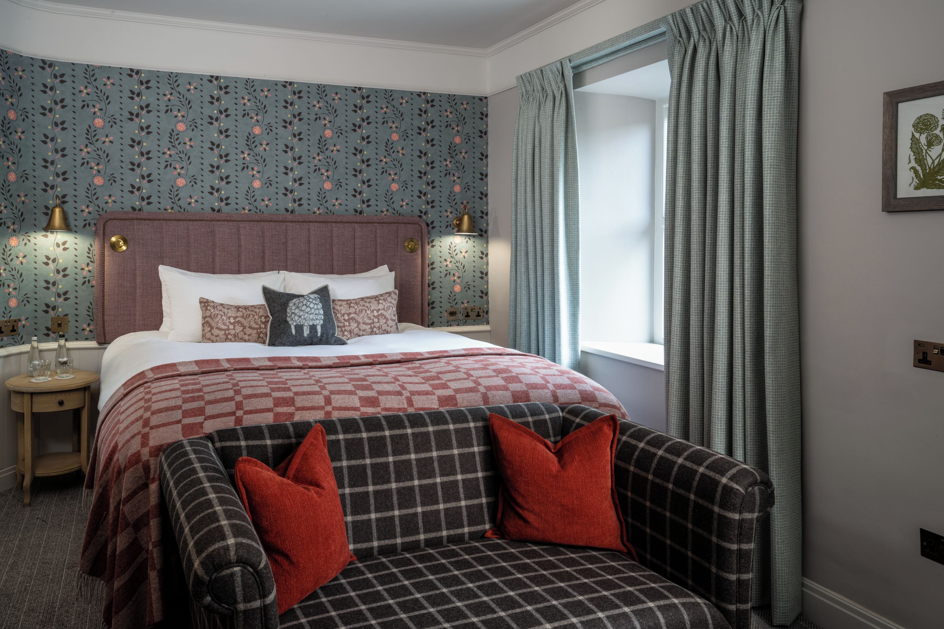 A neatly arranged bedroom with a bed, bedside lamps, a checkered sofa, and a window with curtains.