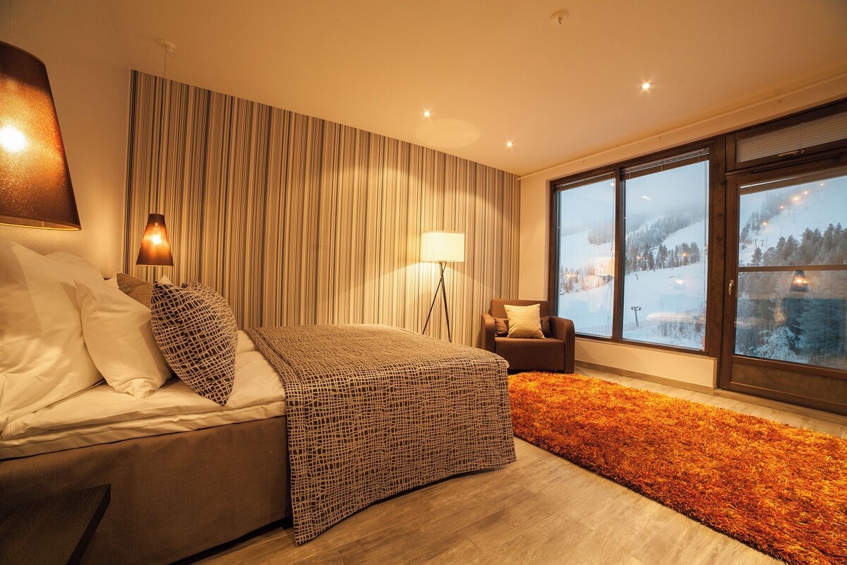 A modern bedroom with a large bed, a standing lamp, a chair, and a view of a snowy landscape through a large window.