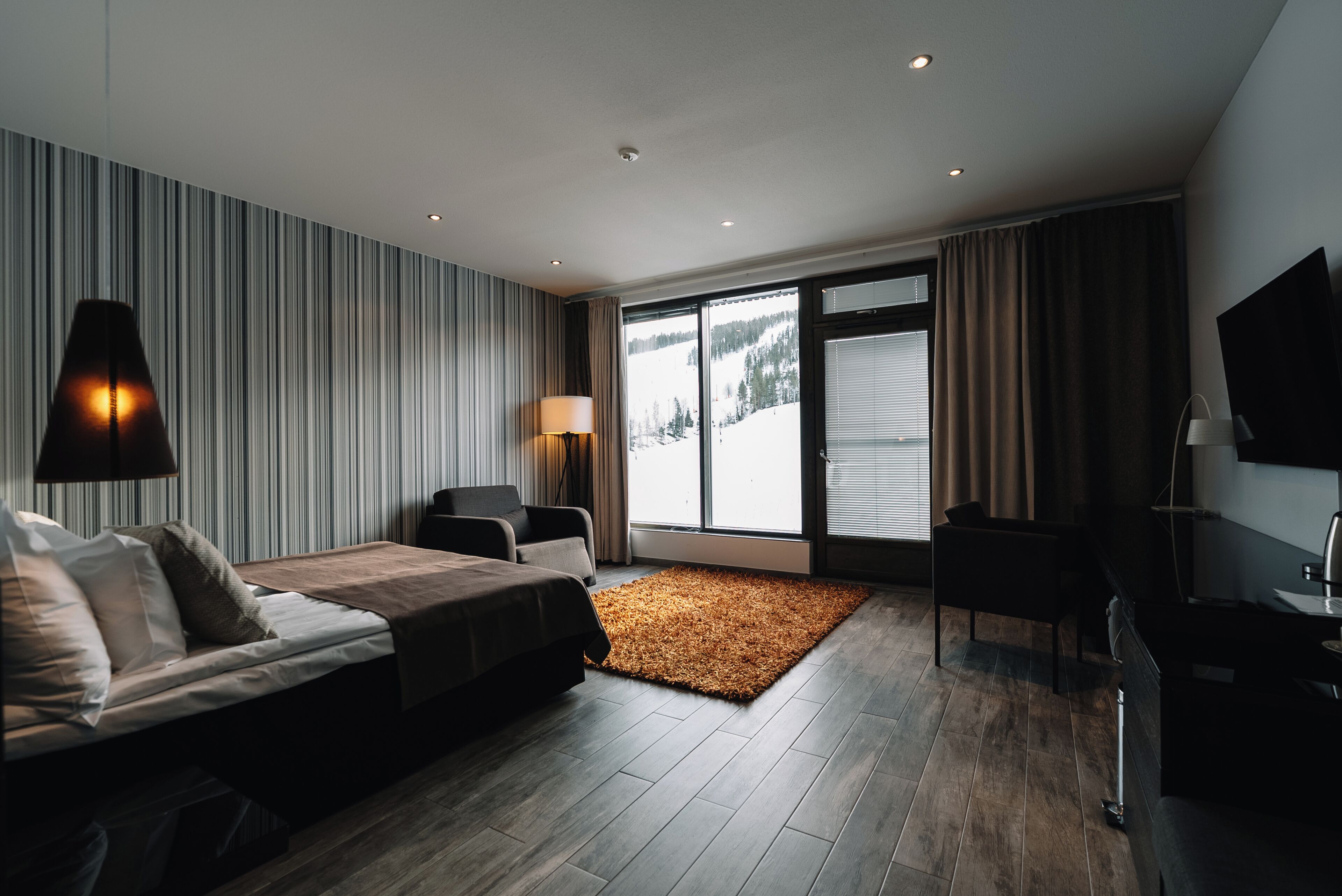 Moderni hotellihuone, jossa on sänky, työpöytä, tuoli, suuri ikkuna, josta avautuu näkymä lumiseen maisemaan, sekä upotetuilla valaisimilla varustettu katto.