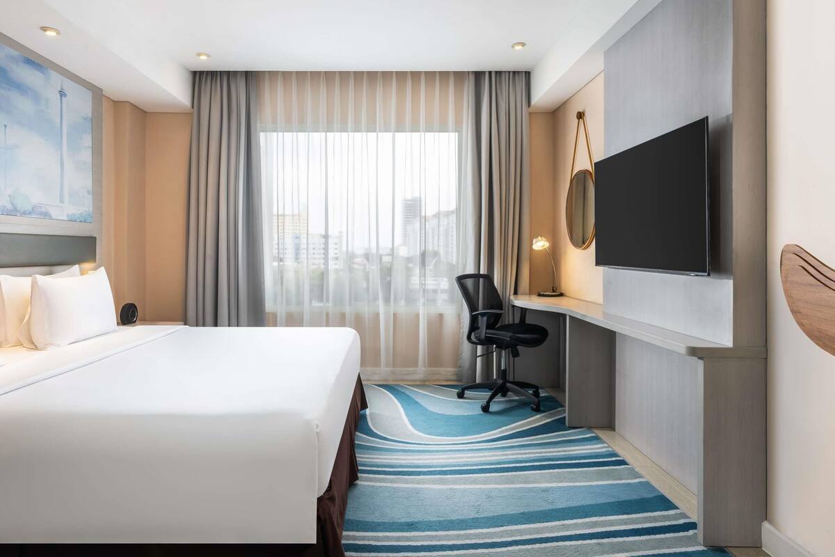 Habitación de hotel con una cama grande, un escritorio con silla, un televisor de pantalla plana y un ventanal con cortinas.