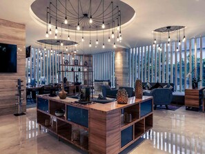 A modern living room with a central wooden island, a TV mounted on the wall, and a large window with vertical blinds. - Mercure Jakarta Cikini (Jakarta)