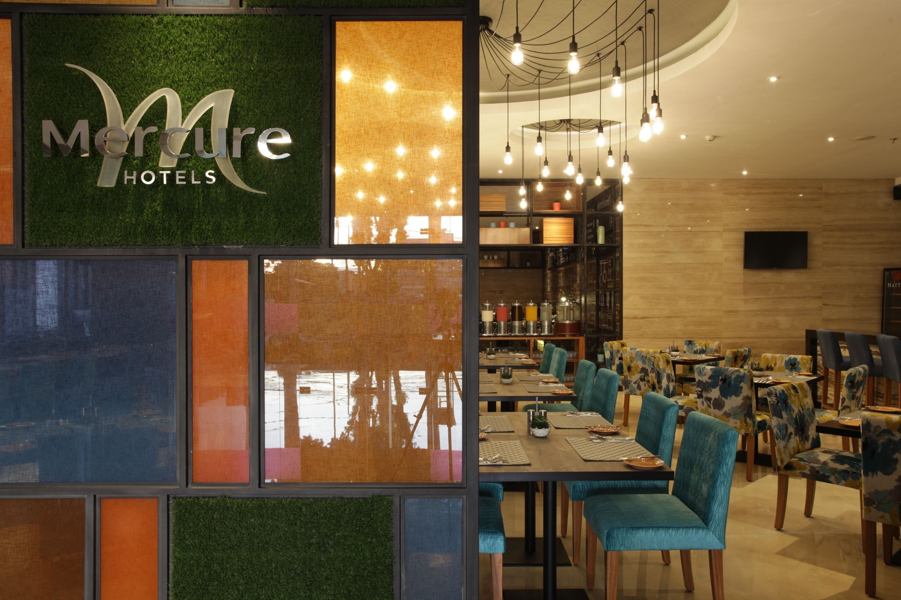 A restaurant area with a green wall displaying the Mercure Hotels logo, wooden tables, and teal upholstered chairs.