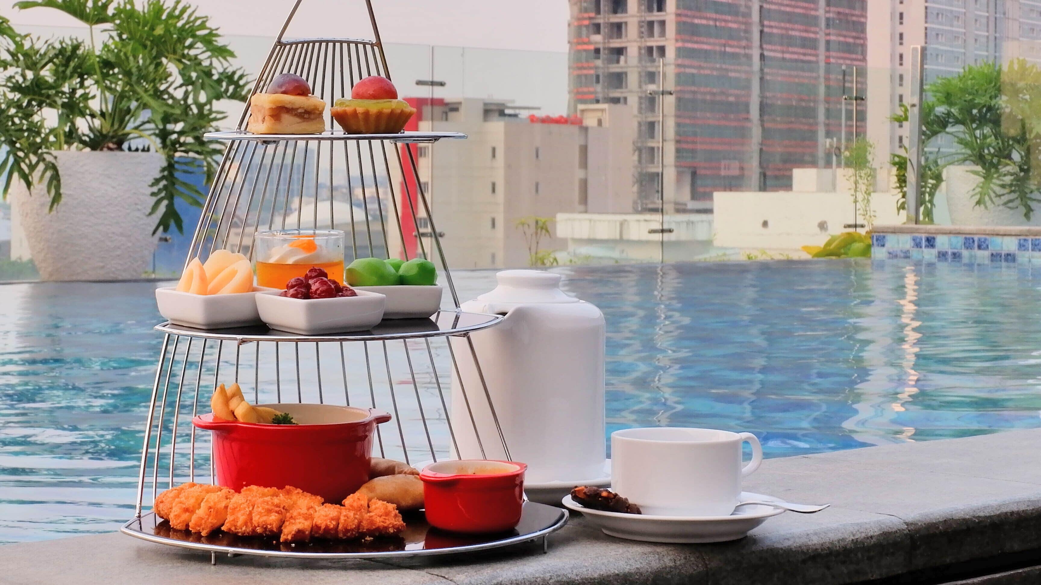 A poolside breakfast setup with a tiered tray holding various fruits, pastries, and a cup of coffee.
