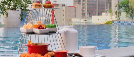A poolside breakfast setup with a tiered tray holding various fruits, pastries, and a cup of coffee.