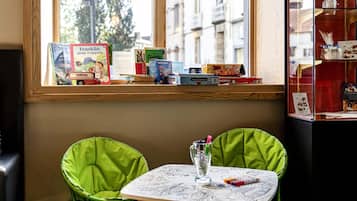 Une pièce avec une fenêtre donnant sur le paysage urbain, une table avec du matériel de dessin et une étagère remplie de livres et de jeux.