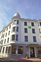 Un edificio blanco con numerosas ventanas y balcones, una estructura en forma de cúpula en la parte superior izquierda y un cartel que dice "ibis Styles" en el lado derecho.