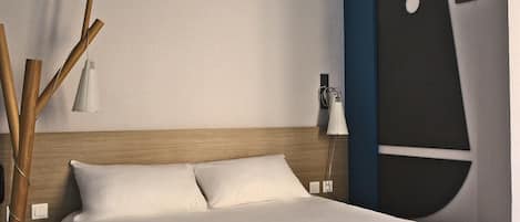 Una habitación de hotel con cabecera de madera, cama con ropa de cama blanca, mesita de noche con lámpara y una obra de arte en la pared.