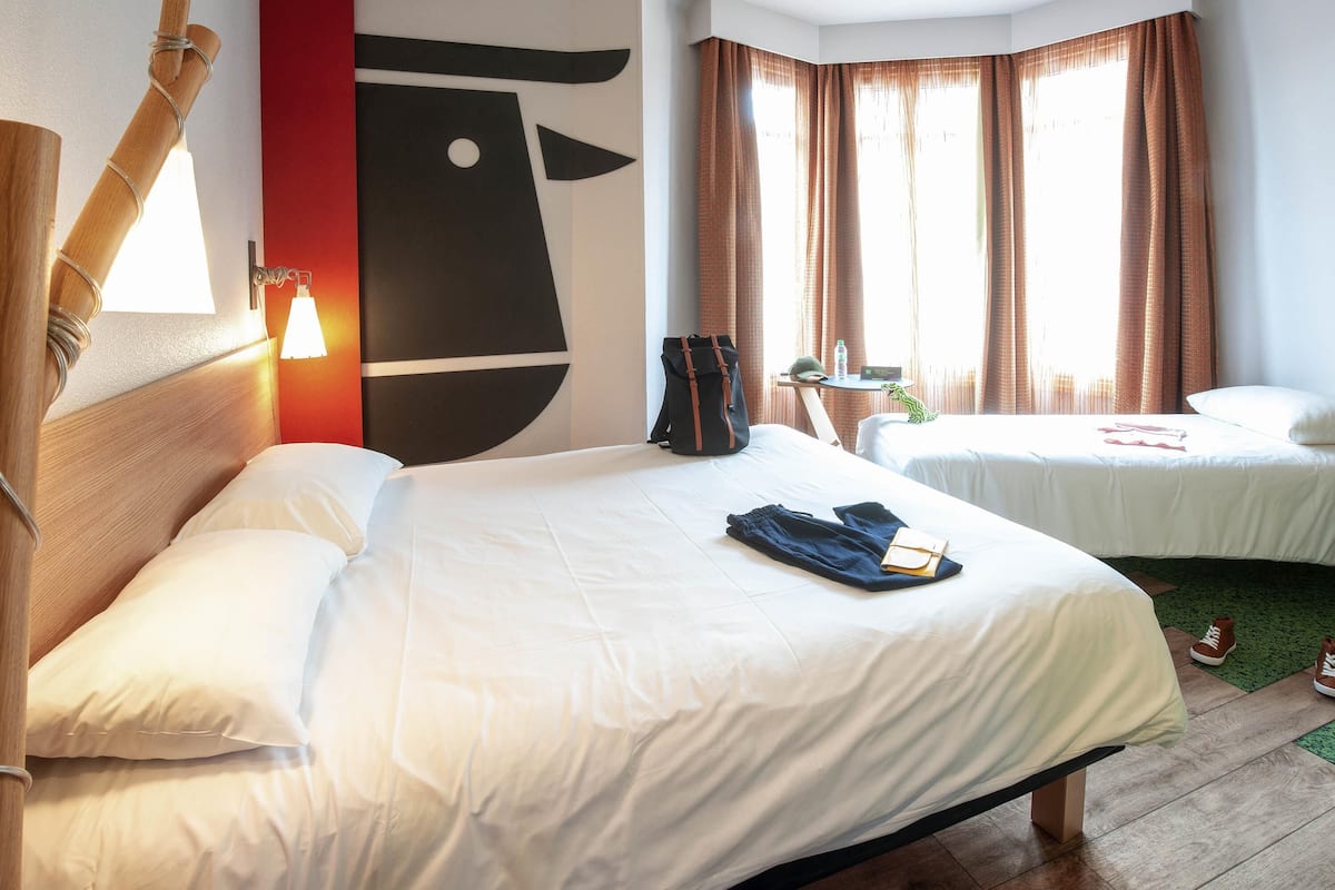 Een hotelkamer met twee bedden, een houten hoofdeinde, een kunstwerk aan de muur, een nachtkastje en een raam met gordijnen.