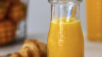 A glass bottle of orange juice with a straw, a croissant, and a blurred background with a basket of oranges.