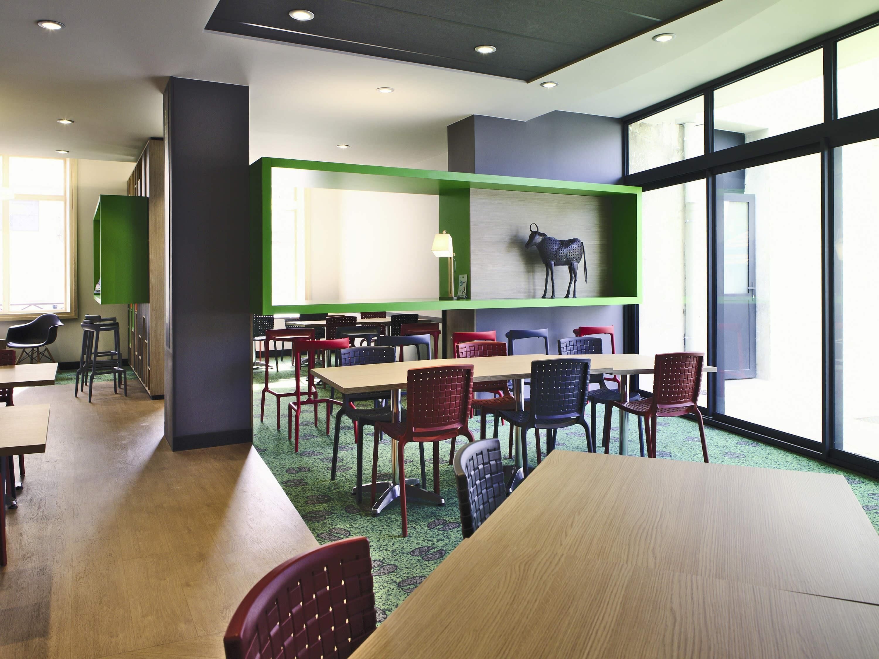 Un comedor moderno con varias mesas y sillas, una pared de acento verde y un ventanal que proporciona luz natural.