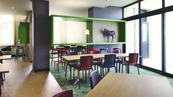 A modern dining area with multiple tables and chairs, a green accent wall, and a large window providing natural light.
